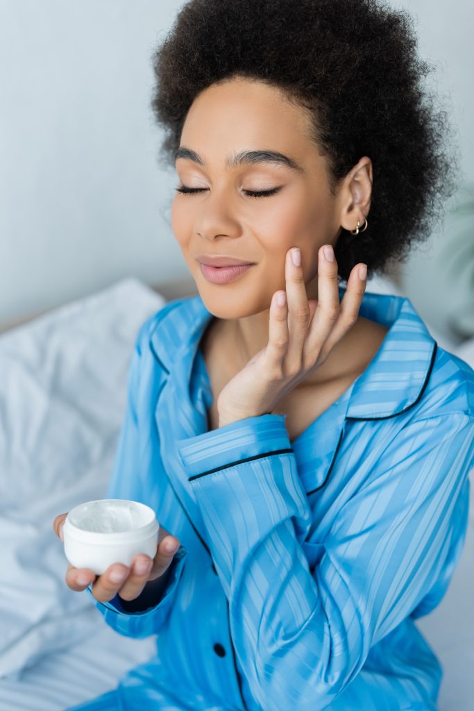 smiling african american woman in pajamas holding container and applying face cream in bedroom