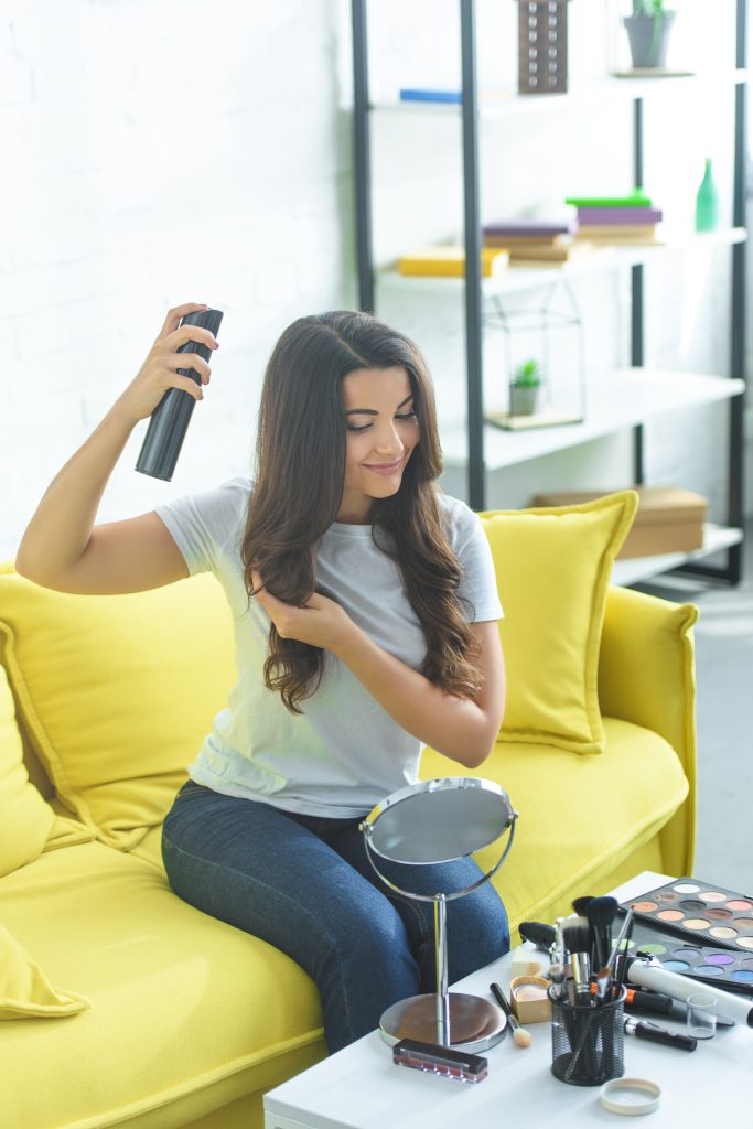 smiling woman applying hair dry shampoo spray while sitting on sofa at home. When to Avoid Dry Shampoo