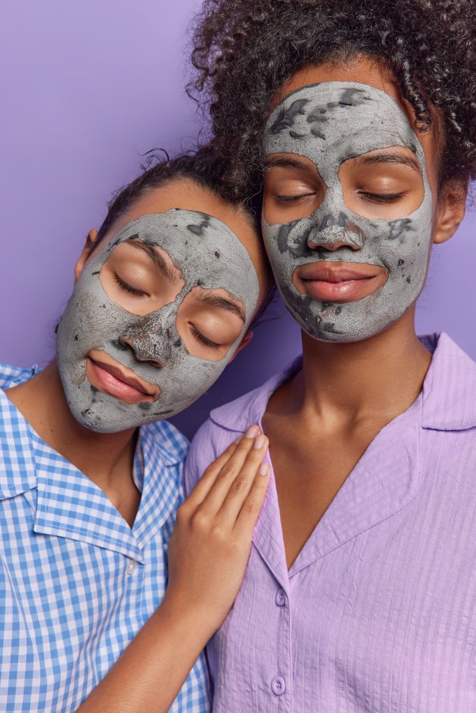 Night Skincare for Different Skin Types. Vertical image of calm young women use cosmetology product apply nourishing grey clay masks on face keep eyes closed dressed in pajamas have calm expressions isolated over purple background..