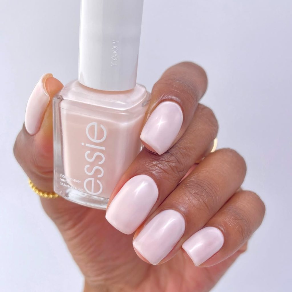 A faceless woman with light pink painted nails holding an Essie nail polish bottle in the same shade. Essie Nail Lacquer Shades.