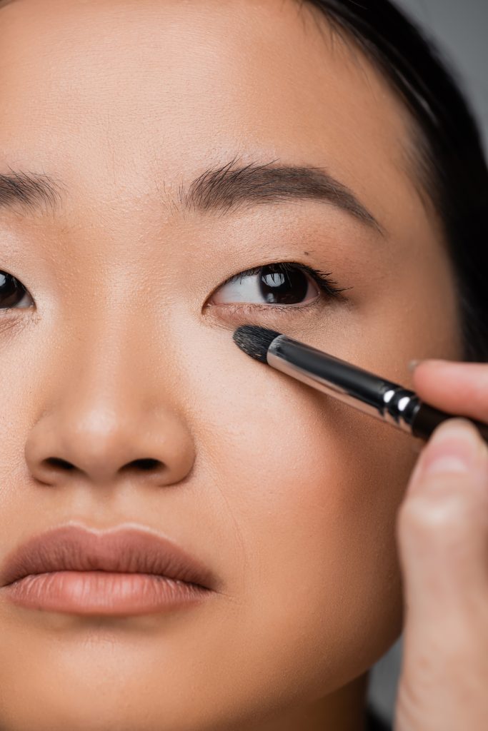 close up view of cropped asian woman with makeup foundation applying eye shadow isolated on grey. Makeup Hacks for Dark Circles. 