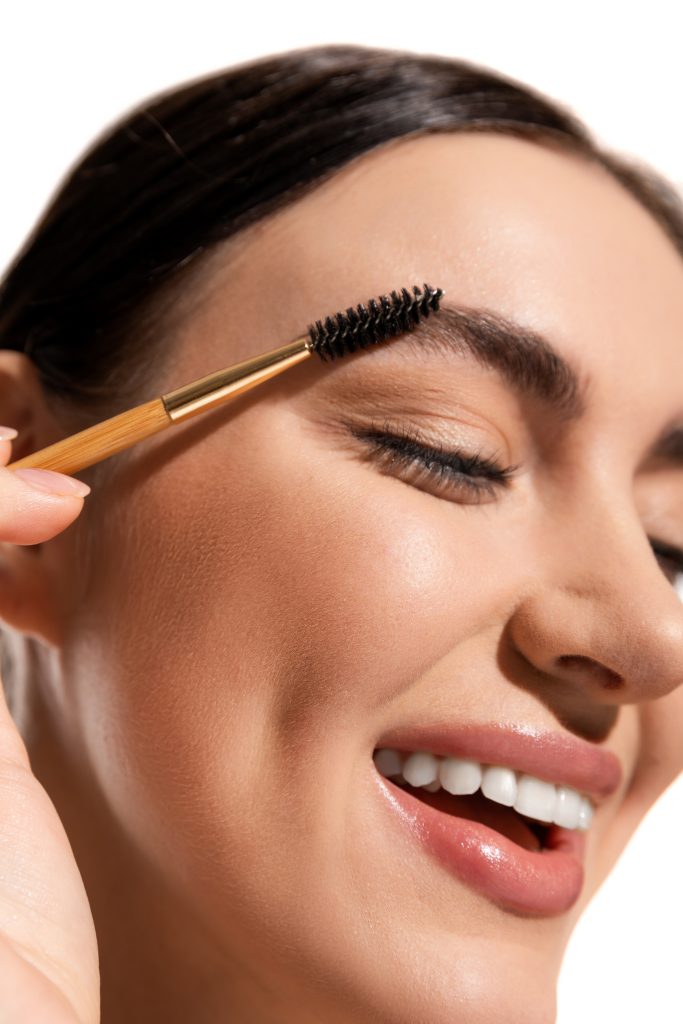 close up view of satisfied young woman styling eyebrows with brush and gel isolated on white