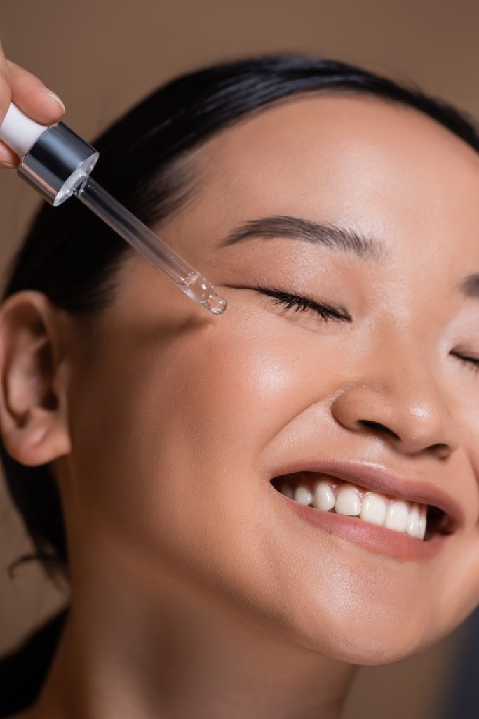 Close up view of smiling asian woman applying cosmetic serum on face isolated on brown. Applying Lash Serum.