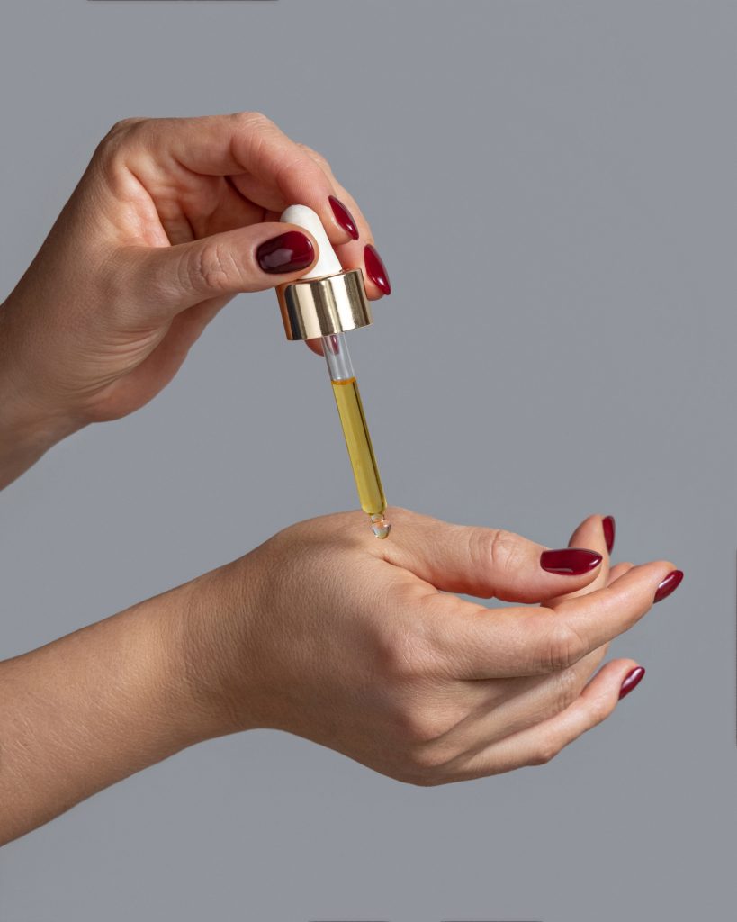 Close up of female hands dropping essential oil on a finger and holding a pipette on other hand against grey background. Healthy hand skin and nails