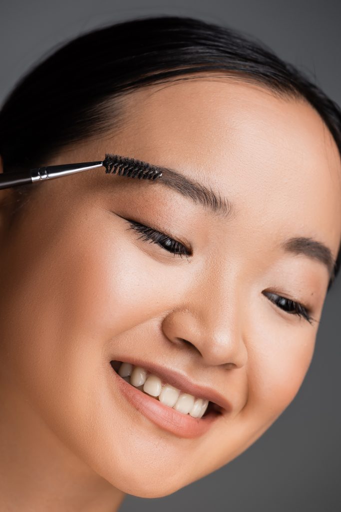 Brow Lamination: The Secret to Gorgeous Arches! portrait of cheerful asian woman with natural makeup brushing eyebrow isolated on grey