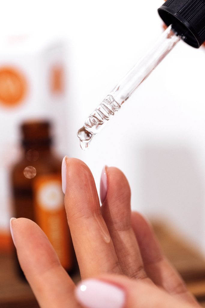 Extreme close up of womans hand and pipette. Transperent drop of essence, oil or gel drips from the pipette. Blurred brown glass serum bottle on the background. Concept of natural organic cosmetics