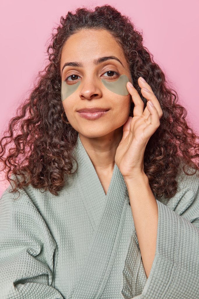 Vertical shot of serious curly haired woman applies hydrogel patches under eyes to reduce wrinkles takes care of facial skin dressed in bathrobe isolated over pink background. Beauty concept. Evening Eye Care Routine.