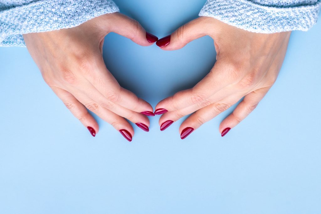 Woman's hands making heart symbol. Female hands in blue knitted sweater fabric with beautiful glossy manicure - burgundy, dark red color nails on blue background with copy space