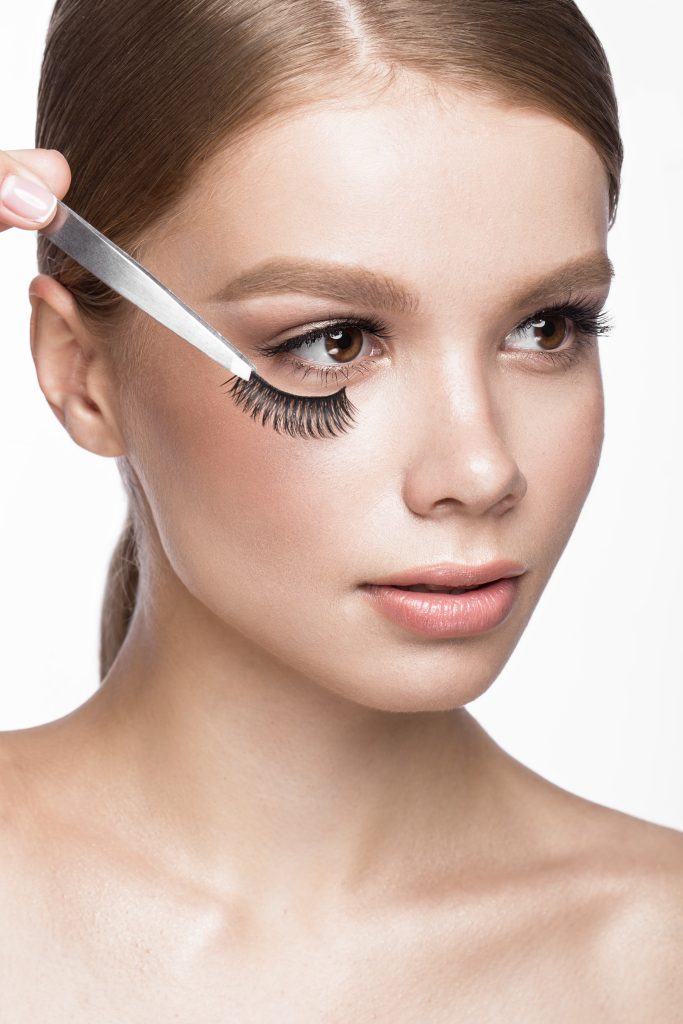 Beautiful young girl with a light natural make-up, Eyebrow Tweezers and False eyelashes . Beauty face. Picture taken in the studio on a white background. Applying False Lashes.