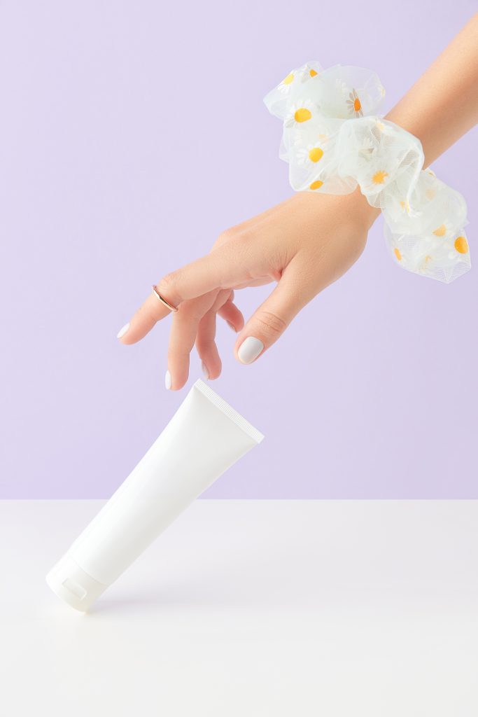 Woman's hand touch white bottle tube on lilac background. Self care beauty treatment concept. Care products bath accessories.