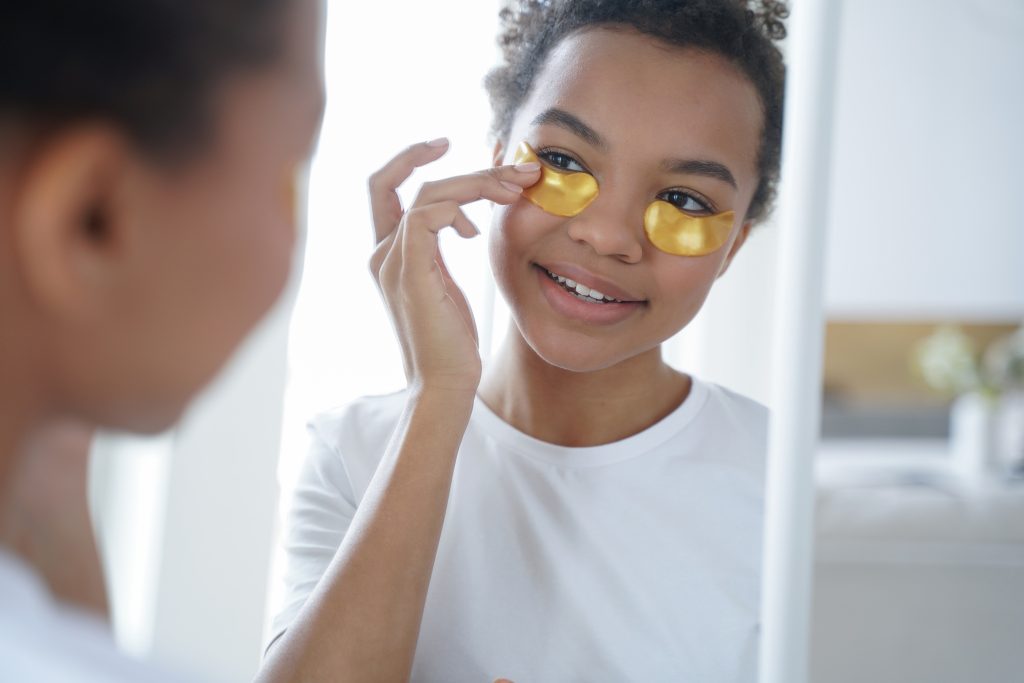 Young teen biracial girl applying moisturizing under eye patches look at mirror. Happy african american woman using gold hydrogel pads reducing facial puffiness, enjoy daily skincare routine. Expert Tips for Dark Circles.