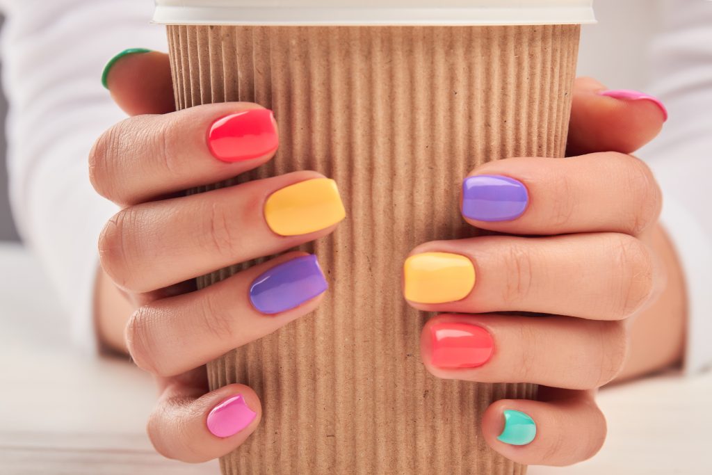 Cardboard cup in female hands close up. Woman hands with multicolored summer manicure holding disposable cup close up. Stylish summer manicure.