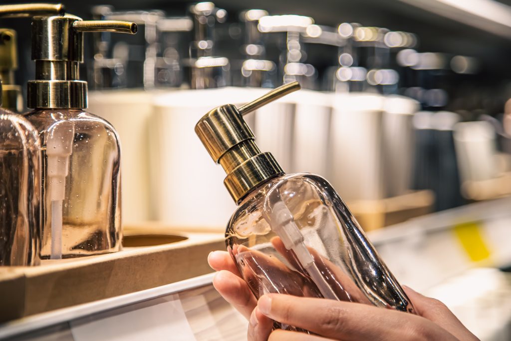 Close up, a dispenser for liquid soap in female hands. Shelves with bathroom accessories in the background. The concept of shopping and sale.