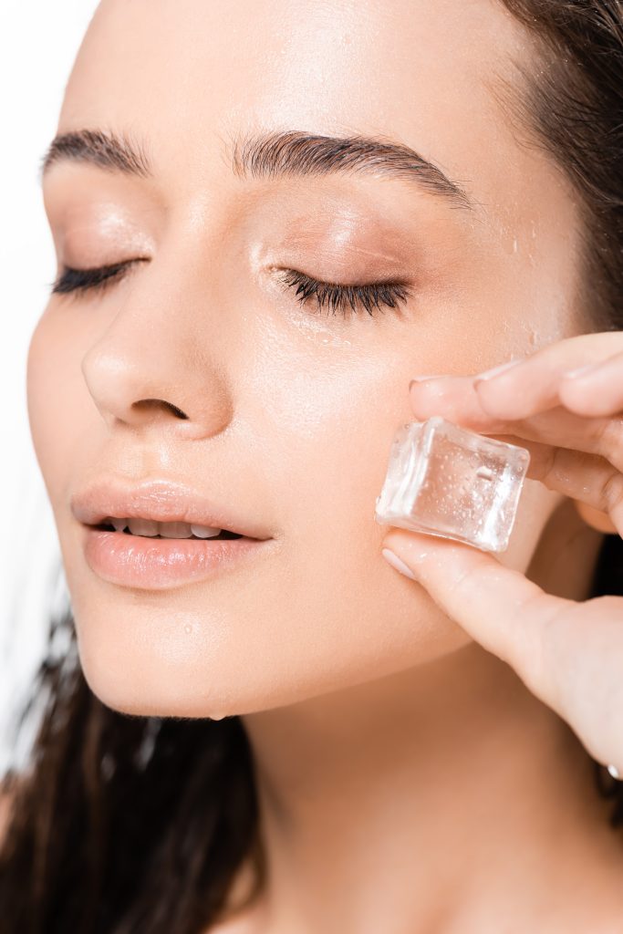 brunette wet young woman with closed eyes and ice cube isolated on white. Skincare Hacks. 