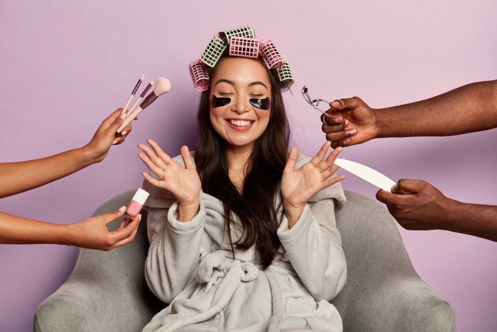 Cheerful brunette mixed race female takes care of face skin, reduces puffiness with eye patches, raises palms from happiness, wears hair curlers, comfortable bathrobe, enjoys beauty treatments. 10 Genius Beauty Hacks That Will Change Your Routine.