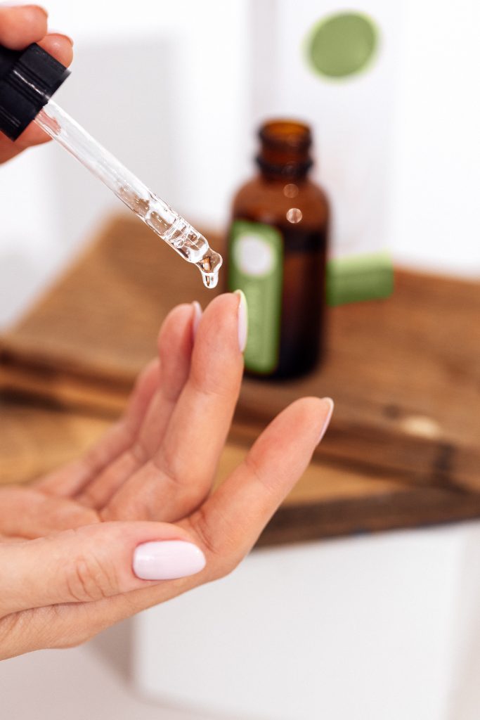 Extreme close up of womans hand and pipette. Transperent drop of essence, oil or gel drips from the pipette. Blurred brown glass serum bottle on the background. Concept of natural organic cosmeti