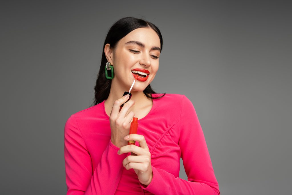 The Best Matte Lipsticks That Stay All Day. gorgeous woman with brunette hair and trendy earrings posing in magenta dress while applying red lip.