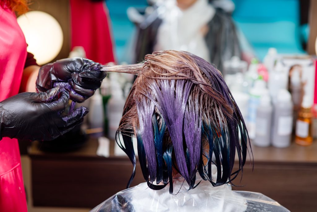 hairdresser thoroughly dyeing hair of female client while she is sitting in chair in beauty salon, top view, close up. Refreshing and Boosting Hair Color Between Treatments.