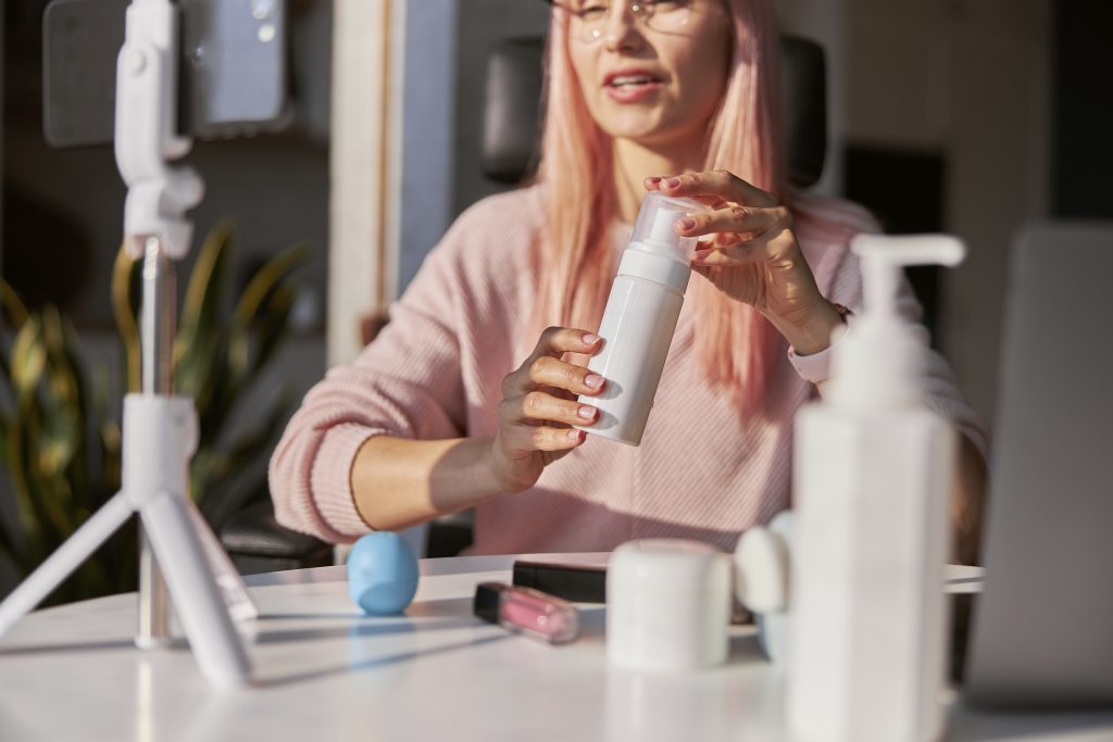 Pretty young woman beauty blogger with pink hair shows bottle of cosmetic product shooting new tutorial at table in sunny room closeup
