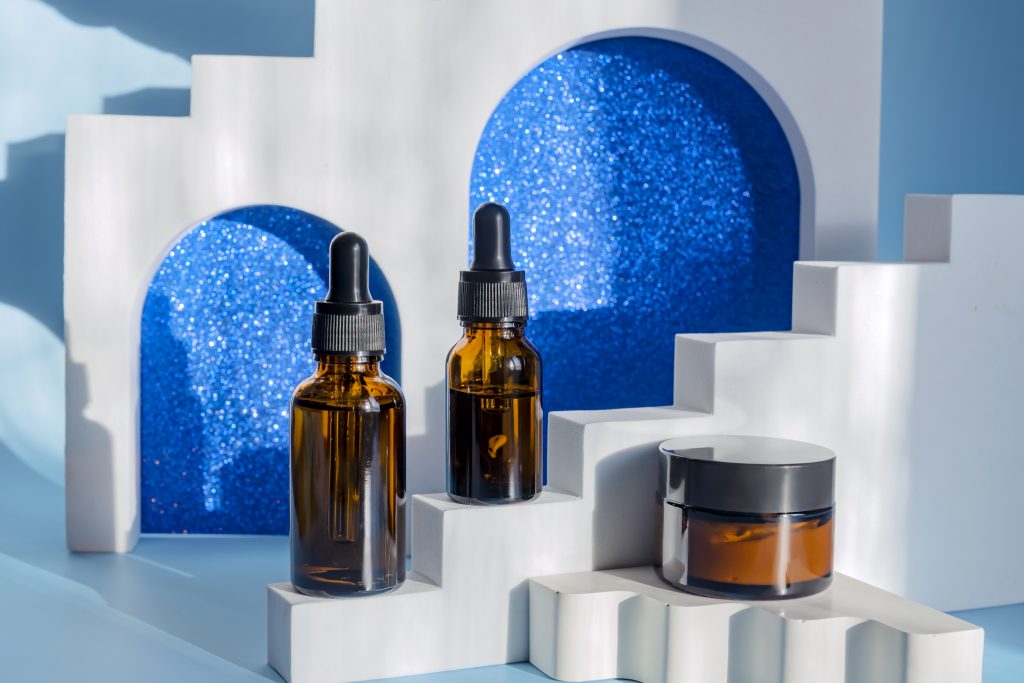 Trendy shoot of cosmetic packaging. Amber glass dropper bottles and cream jar om white podiums, arches and stairs. Skincare festive concept