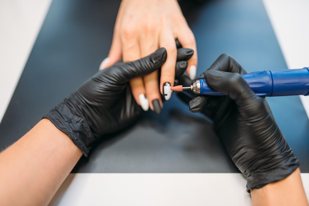 Beautician in gloves holds polishing machine and cleans old varnish from nails of female client, top view, manicure in beauty salon. Manicurist doing hands care cosmetic procedure. Why Polygel Is Dominating the Nail Scene in 2025.