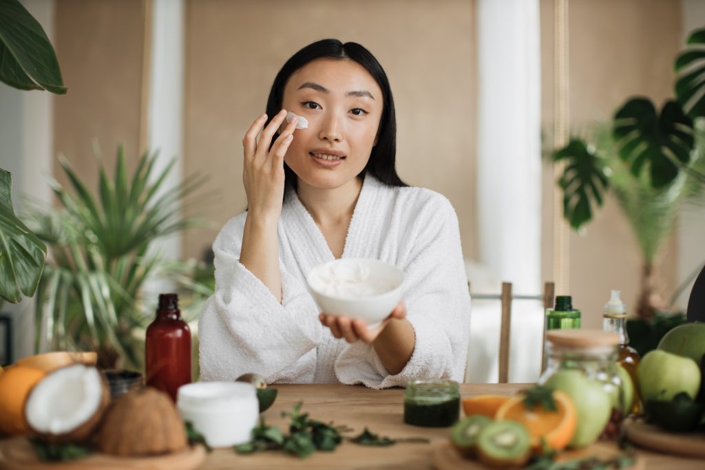 Beautiful asian woman preparing homemade cream with natural ingredients for skin care applying on face sitting at wooden table with various organic fresh vegetables and fruits on light exotic studio