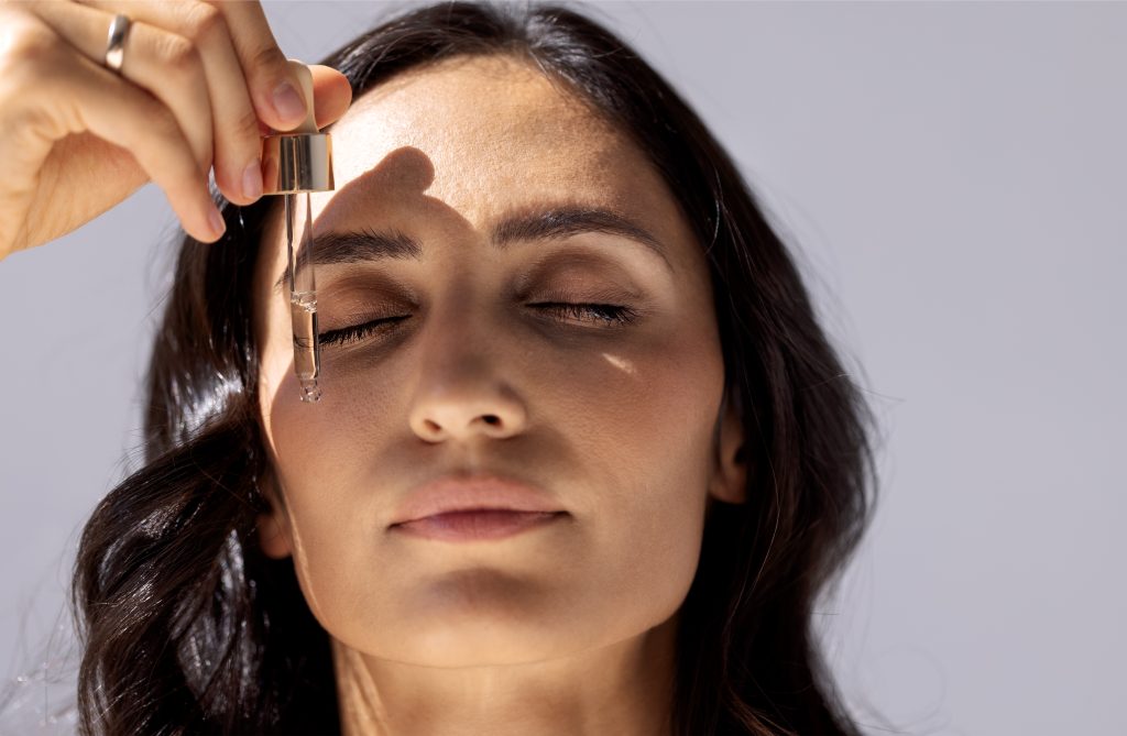 Close-up of charming woman with her eyes closed holding pipette with regenerating serum with hyaluronic acid. Attractive girl with beautiful healthy skin and moisturizing anti-aging facial essence.