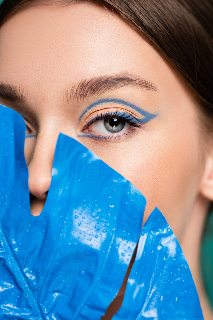 close up view of woman with creative makeup obscuring face with blue leaf