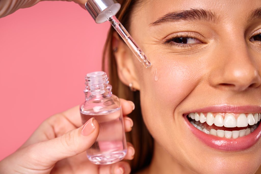 Beautiful woman face, natural clean and fresh glowing skin, applying anti-aging collagen serum. Hands hold serum with active herbal ingredients vitamin C in a dropper, on a pink background. toner