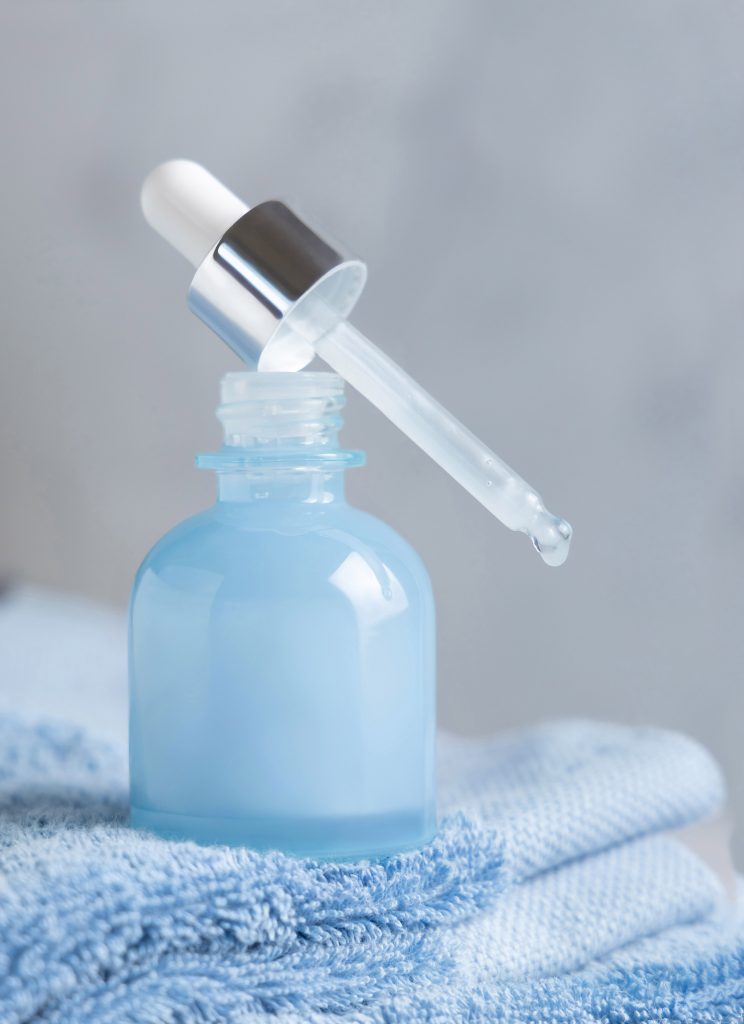 Drop falling from pipette set on opened blue glass dropper bottle on blue towel against grey close up, mockup. Natural serum or essence