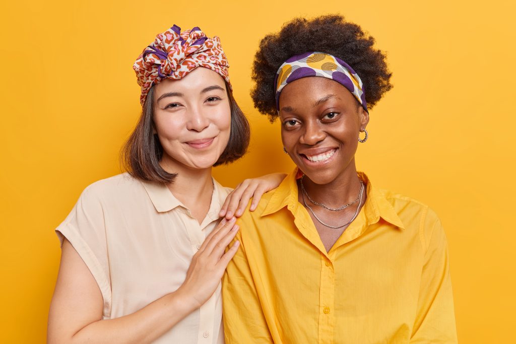 Portrait of cheerful mixed race women smile pleasantly spend free time together missed each other dressed casually isolated over yellow background. Diverse female friends of different nationalities