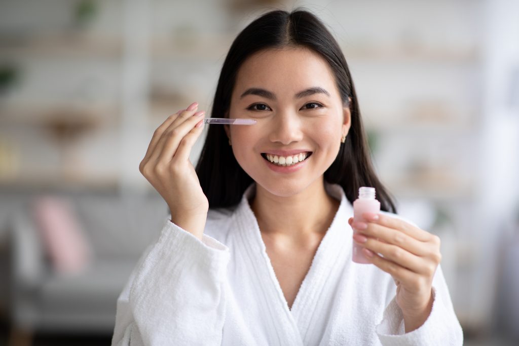 Pretty asian lady applying eye serum and smiling at camera, wearing white bathrobe, holding bottle with beuty product, copy space. Beautiful korean woman using anti-aging eye essence