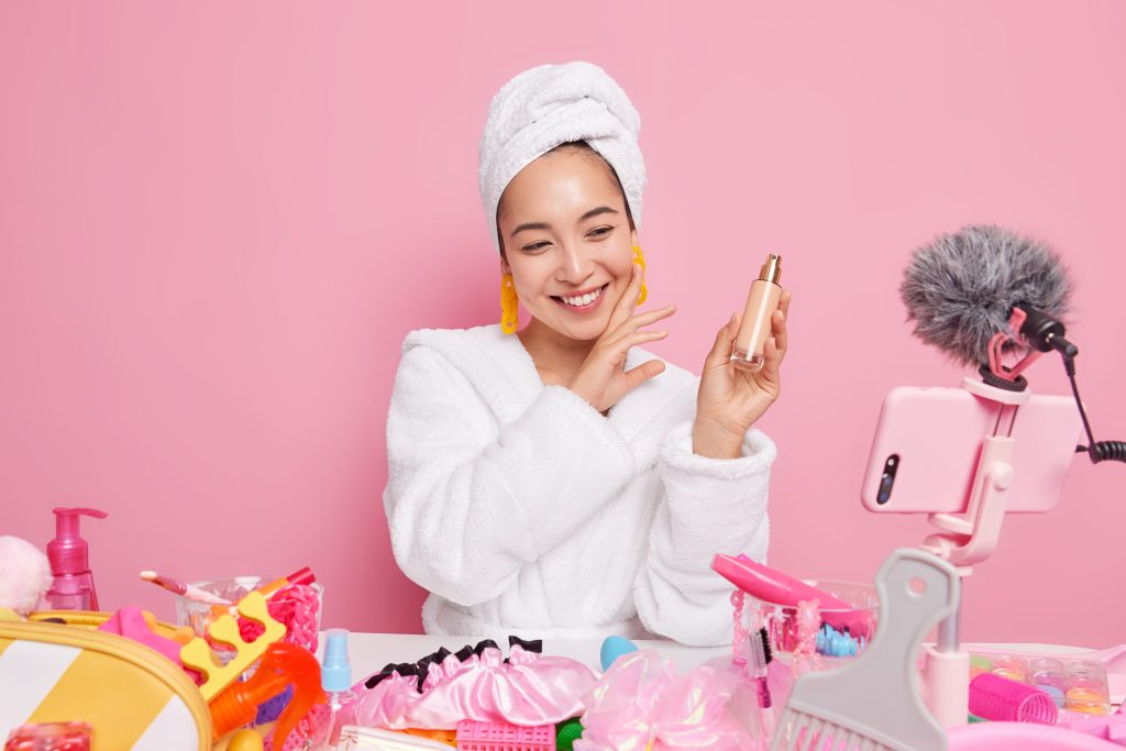 Smiling young Asian woman does cosmetic overview tries new foundation records video blog wears bathrobe towel wrapped over head sits at table wih beauty products isolated over pink background