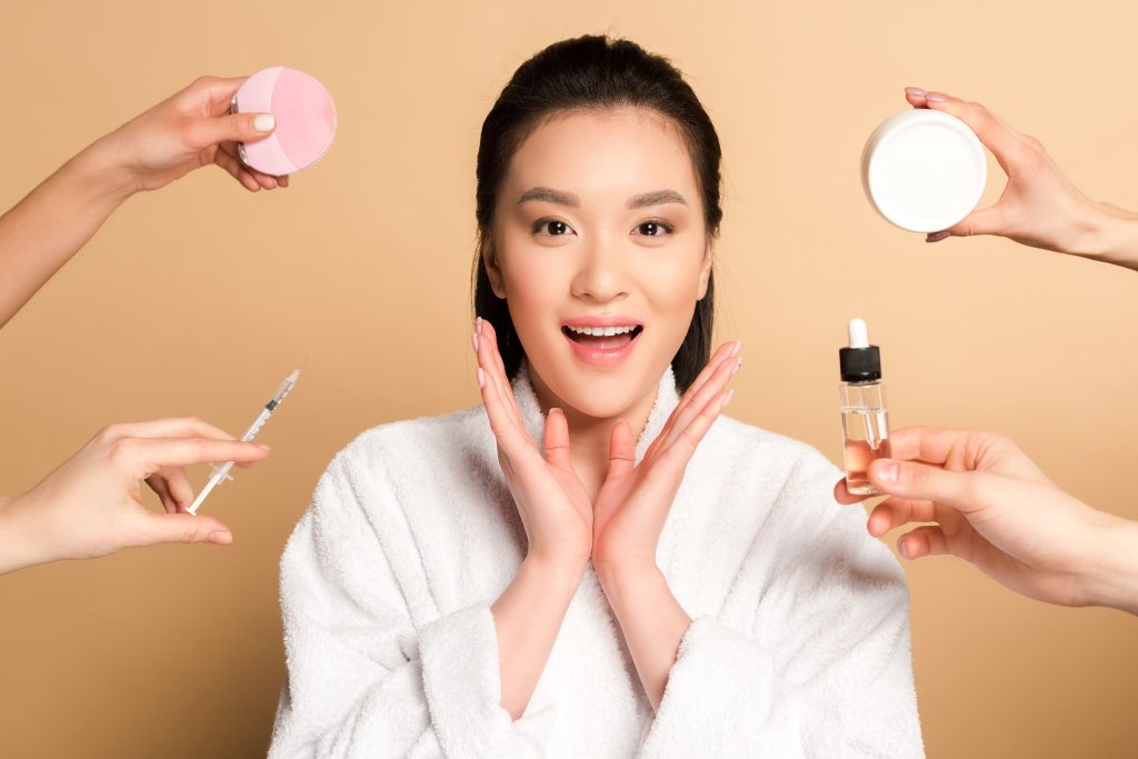 surprised beautiful asian woman in bathrobe near hands with facial cleansing brush, syringe,