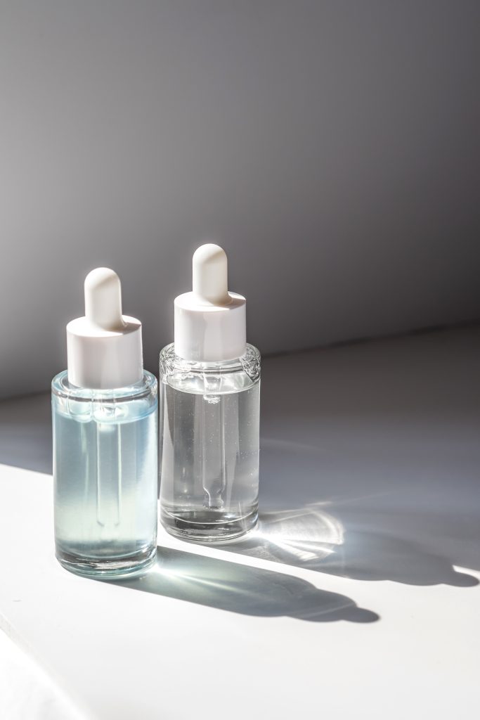 Two laconic bottles of cosmetic liquid in the rays of the harsh sun. A transparent white bottle with a pipette and a transparent blue one.