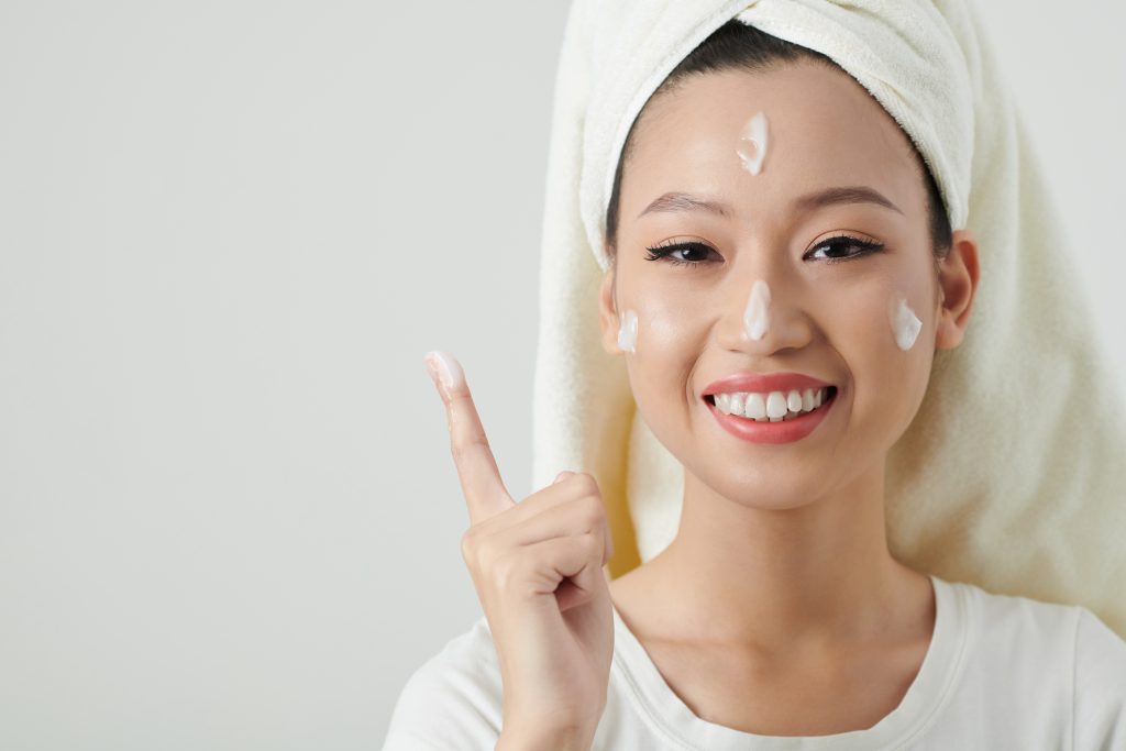 Positive young woman applying face cream to moisturize her skin after shower