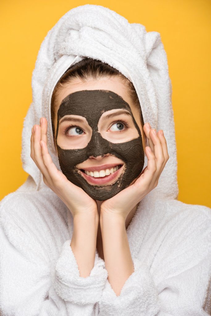 How Black Facial Masks Deliver Clear and Healthy Skin Results.. cheerful girl with facial clay mask and towel on head holding hands near face and looking away