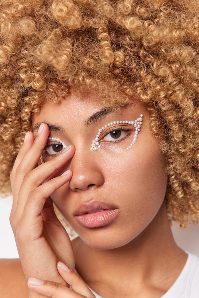 Close up shot of serious calm young gentle woman keeps hand on face looks directly at camera has well cared skin white beads around eye curly bushy hair poses indoor. Natural beauty concept.