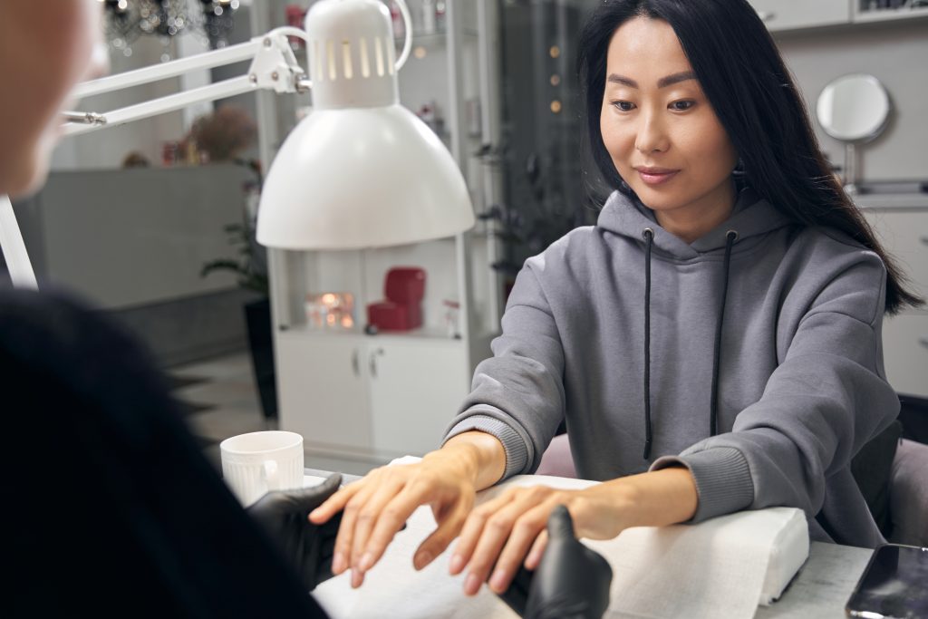 Charming Asian female in grey hoodie receiving a manicure procedure in beauty center