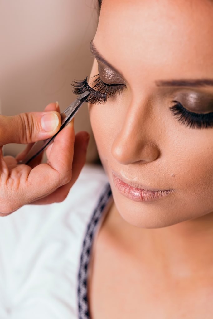 Make up artist working in a make up studio, putting artificial eyelashes on female client's eyelids with a tweezers.