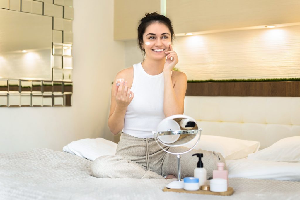 A young caucasian smiling woman is sitting on the bed and doing skin care, mirror and skin care products nearby