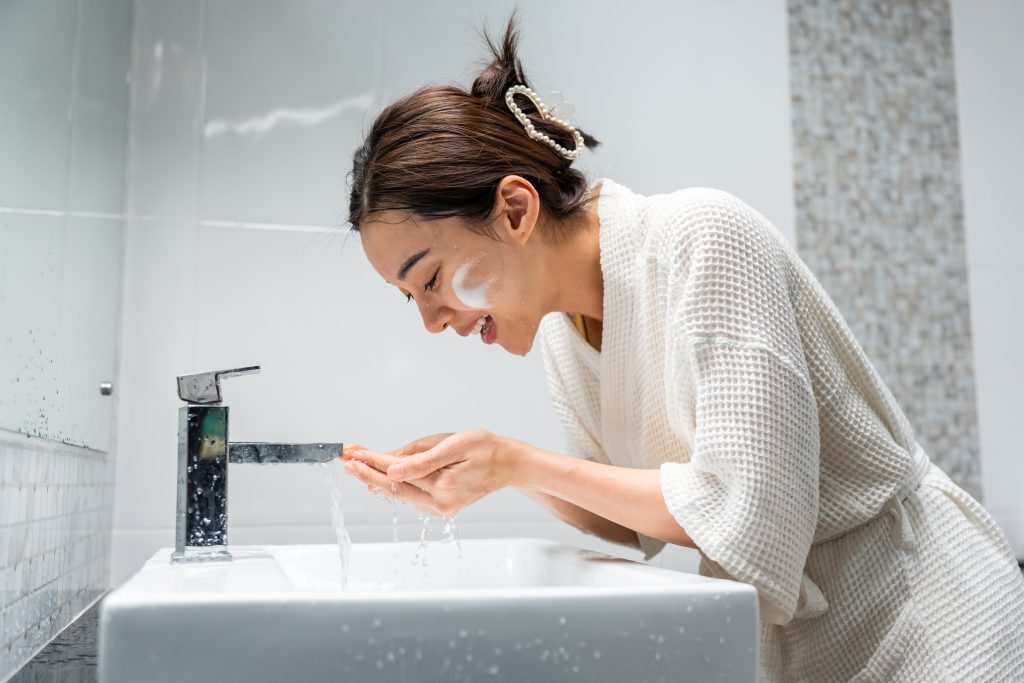 Double Cleansing. Asian beautiful woman washing her clean face with facial foam and water. Attractive female in bathrobe washing face for healthy beauty treatments and skin care then looking at the mirror in bathroom.