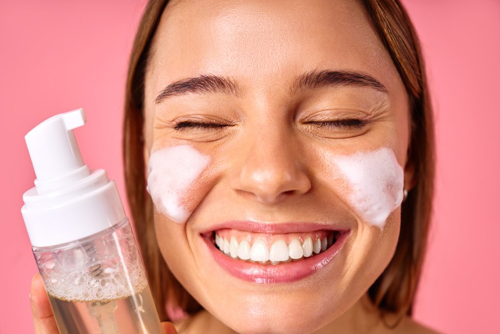Beautiful woman cleansing her face with foam isolated on pink background. Facial skin care, cosmetology, beauty concept.