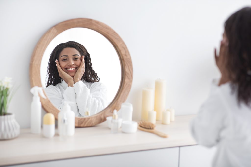 Enjoy result of beauty therapy, spa at home on weekends or free time. Cheerful young african american female rejoices to perfect shiny skin, looks at reflection of round mirror in room interior