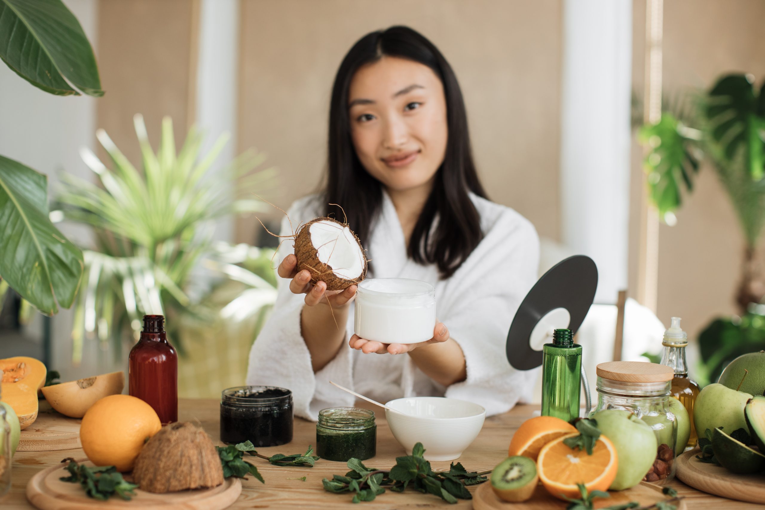 Anti-Aging Ingredients. Anti-Aging skincare Ingredients. Focus on coconut and glass jar, young asian woman holding half of coconut and making homemade cream for healthy skin. A lot of ingredients for homemade cosmetics lying on wooden table.