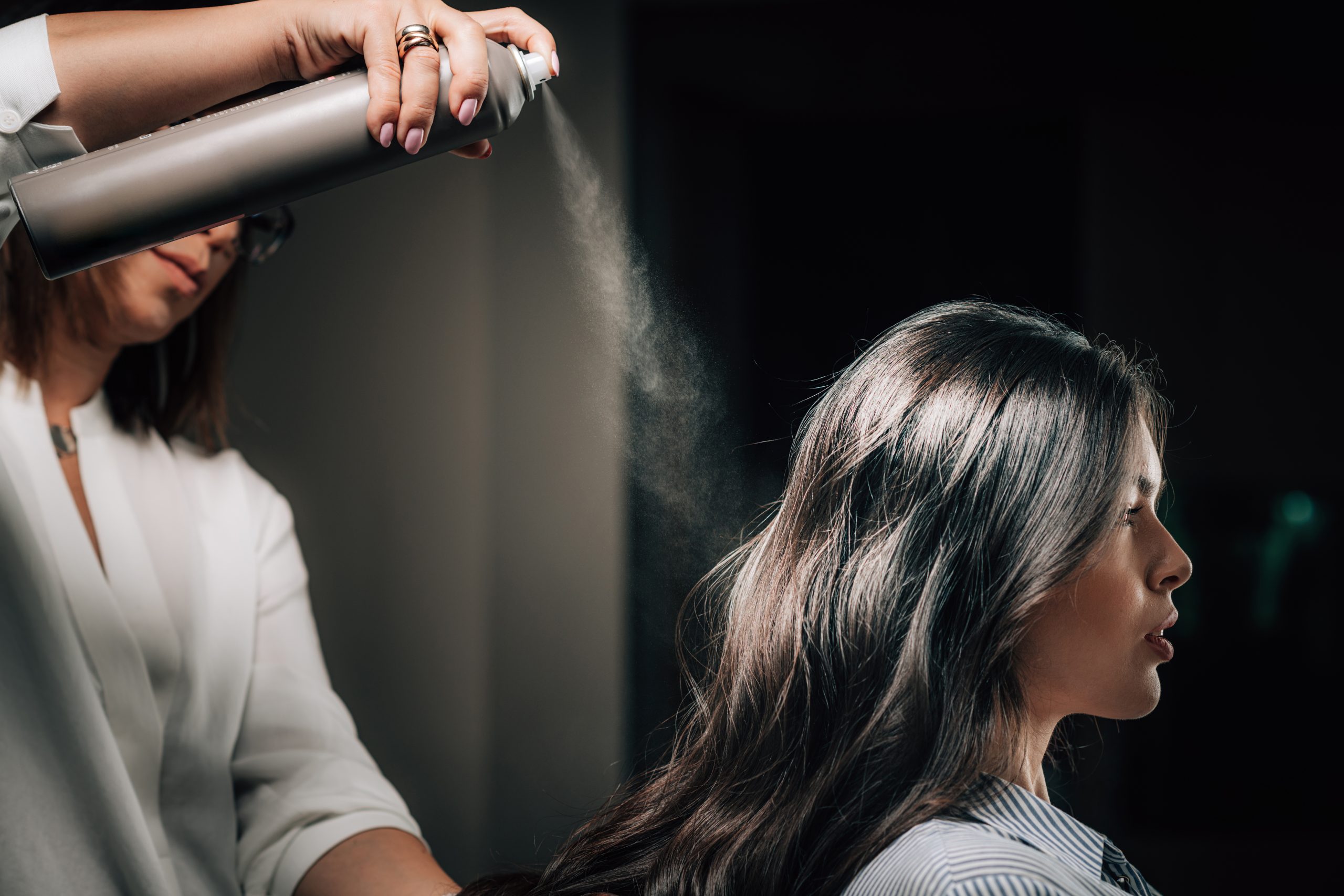 Hair Heat Protectants. Hairdresser spraying woman’s long black hair with hair spray.