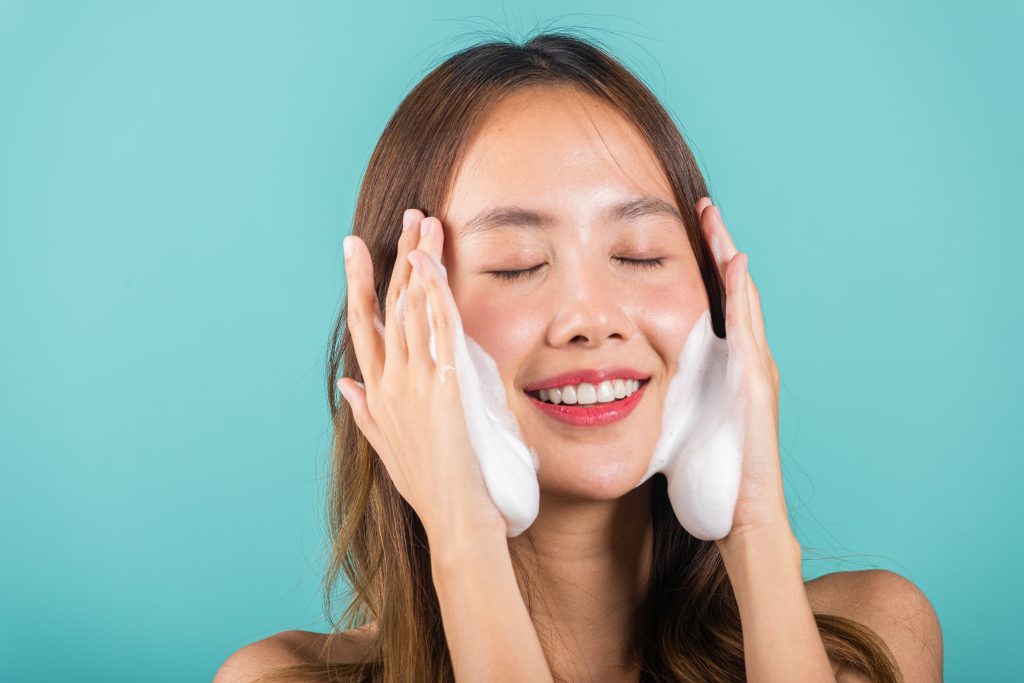 Double Cleansing. Portrait beautiful female smile applies foaming cleanser healthy skin, Asian women face wash exfoliating scrub soap with skincare cleansing, Studio isolated on blue background, skincare daily routine