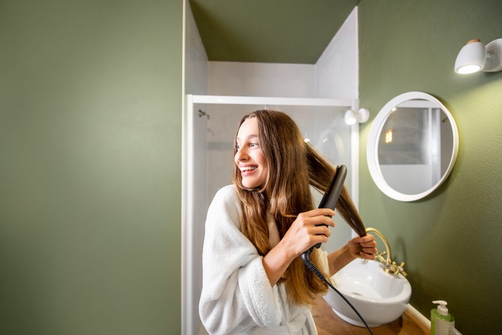 Hair Loss Causes. Young and happy woman in bathrobe straightening hair with straightener in the bathroom.