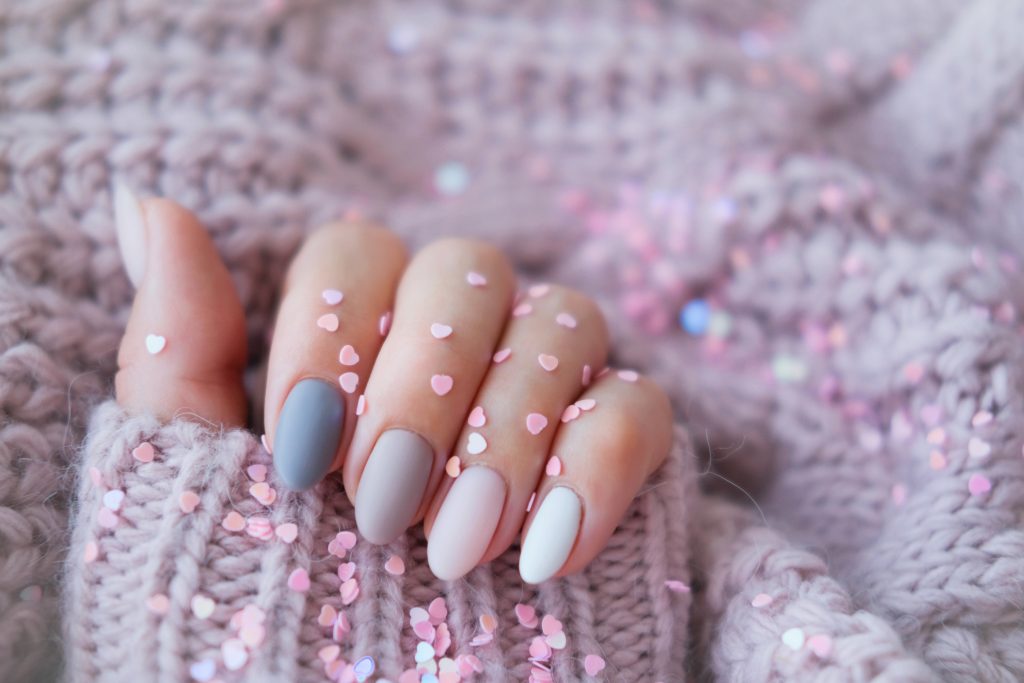 Women's hands with a beautiful matte oval manicure in a warm purple knitted sweater and heart shaped sequins. Winter trend, polish beige nails with gel polish, shellac. Copy space.