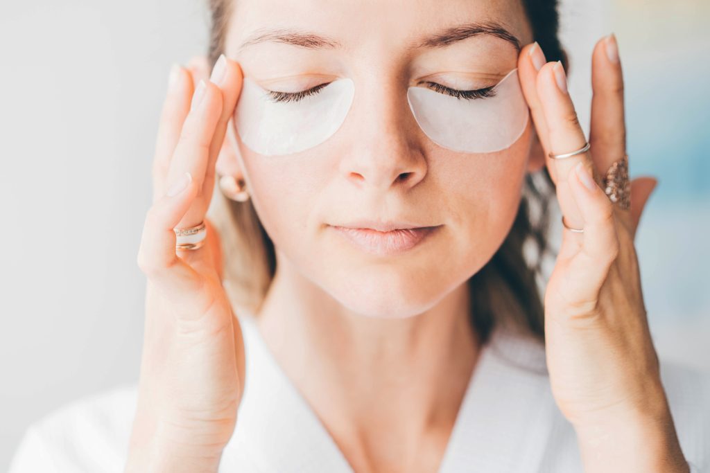 Attractive mature woman in white bathrobe puts anti aging eye patch on face taking care of skin in light modern hotel room close view. Eye Masks